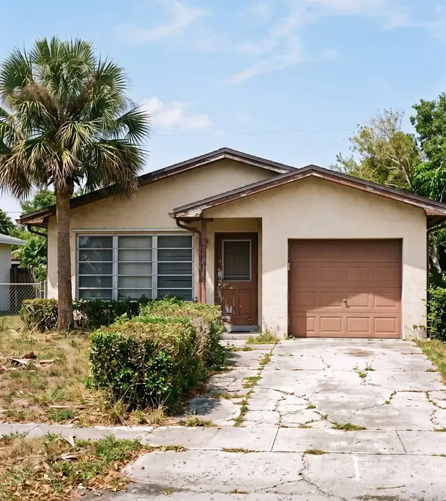 Dated 1970s Florida house with beige stucco, jalousie windows, a brown garage door and a cracked driveway