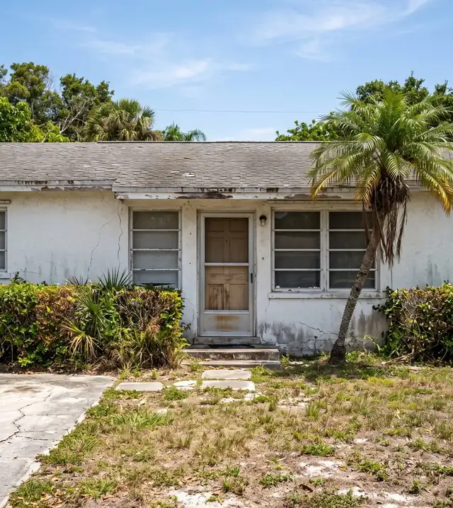 Front of a worn white stucco Florida house with cracked walls, an old screen door and overgrown hedges